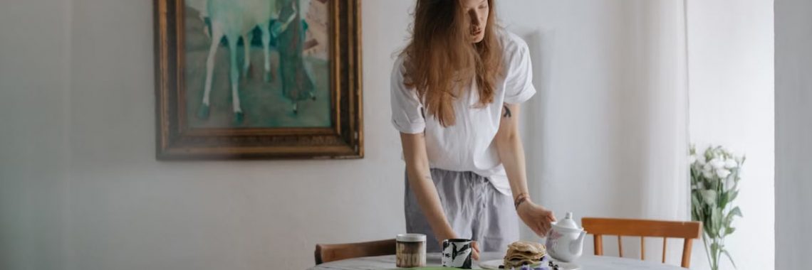 A woman in a casual setting prepares a table with pancakes and a teapot. Artistic interior vibes.